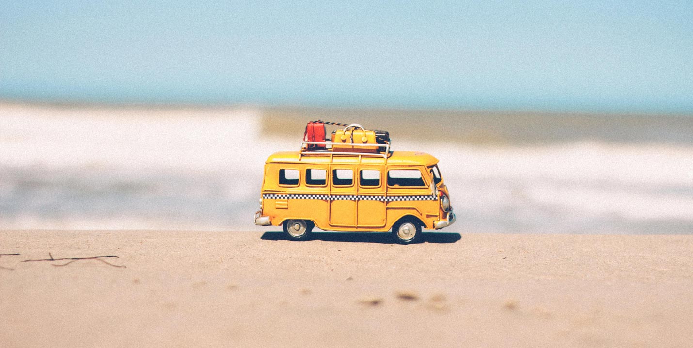 Gelber Spielzeugbus mit Gepäck auf dem Dach vor einem unscharfen Strand und blauem Himmel