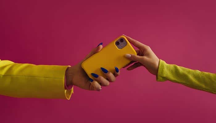 Zwei Hände reichen sich ein gelbes Smartphone vor pinkfarbenem Hintergrund