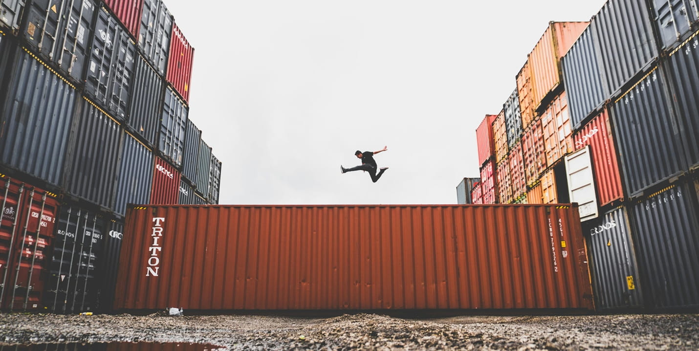 Person springt über einen roten Frachtcontainer zwischen hohen Stapeln aus verschiedenfarbigen Containern