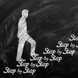 Kreidezeichnung einer Person, die auf einer schwarzen Tafel eine Treppe mit der mehrfachen Aufschrift „Step by Step“ hinaufsteigt