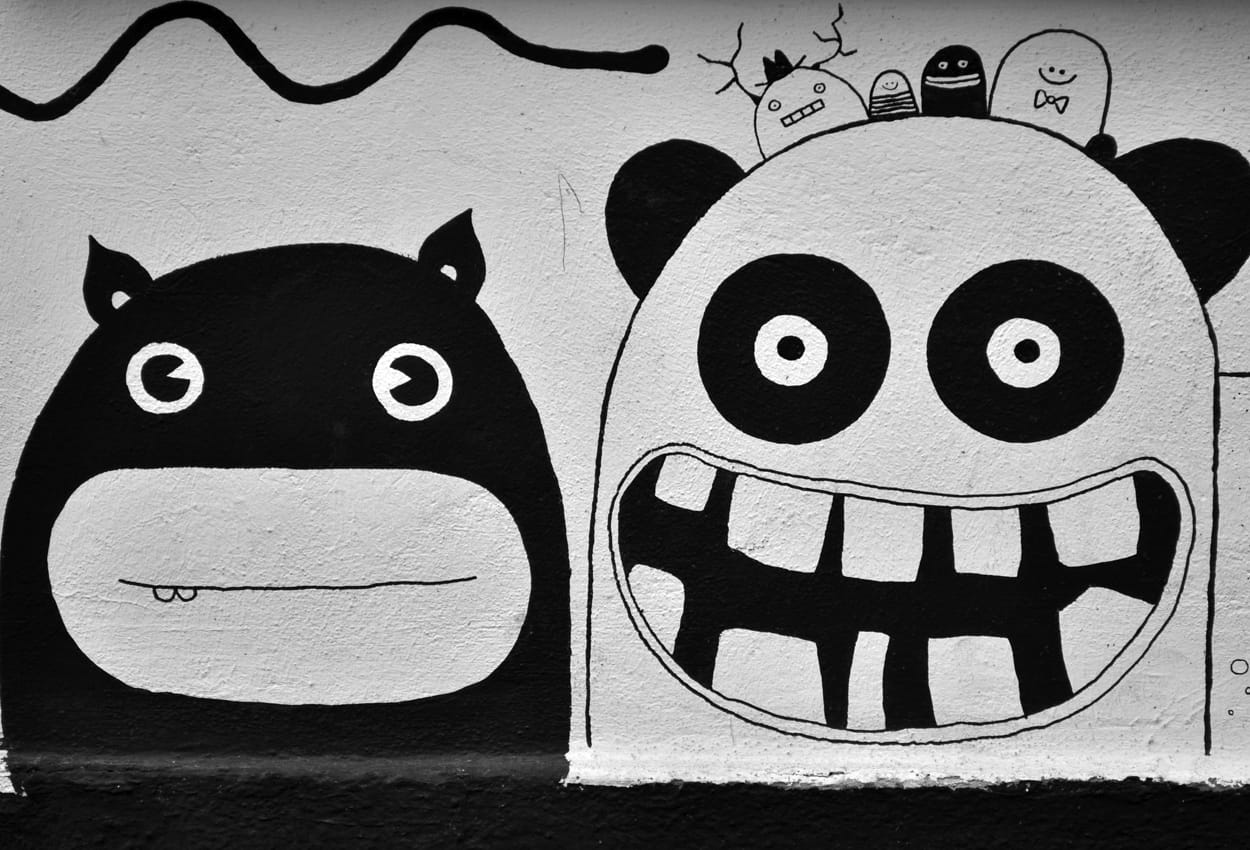 Schwarz-weißes Wandgemälde mit zwei großen, comicartigen Monstergesichtern – links ein rundäugiges Wesen mit geschlossenem Mund, rechts ein grinsendes Wesen mit Zähnen und Ohren, darüber kleinere gezeichnete Figuren und eine geschwungene Linie