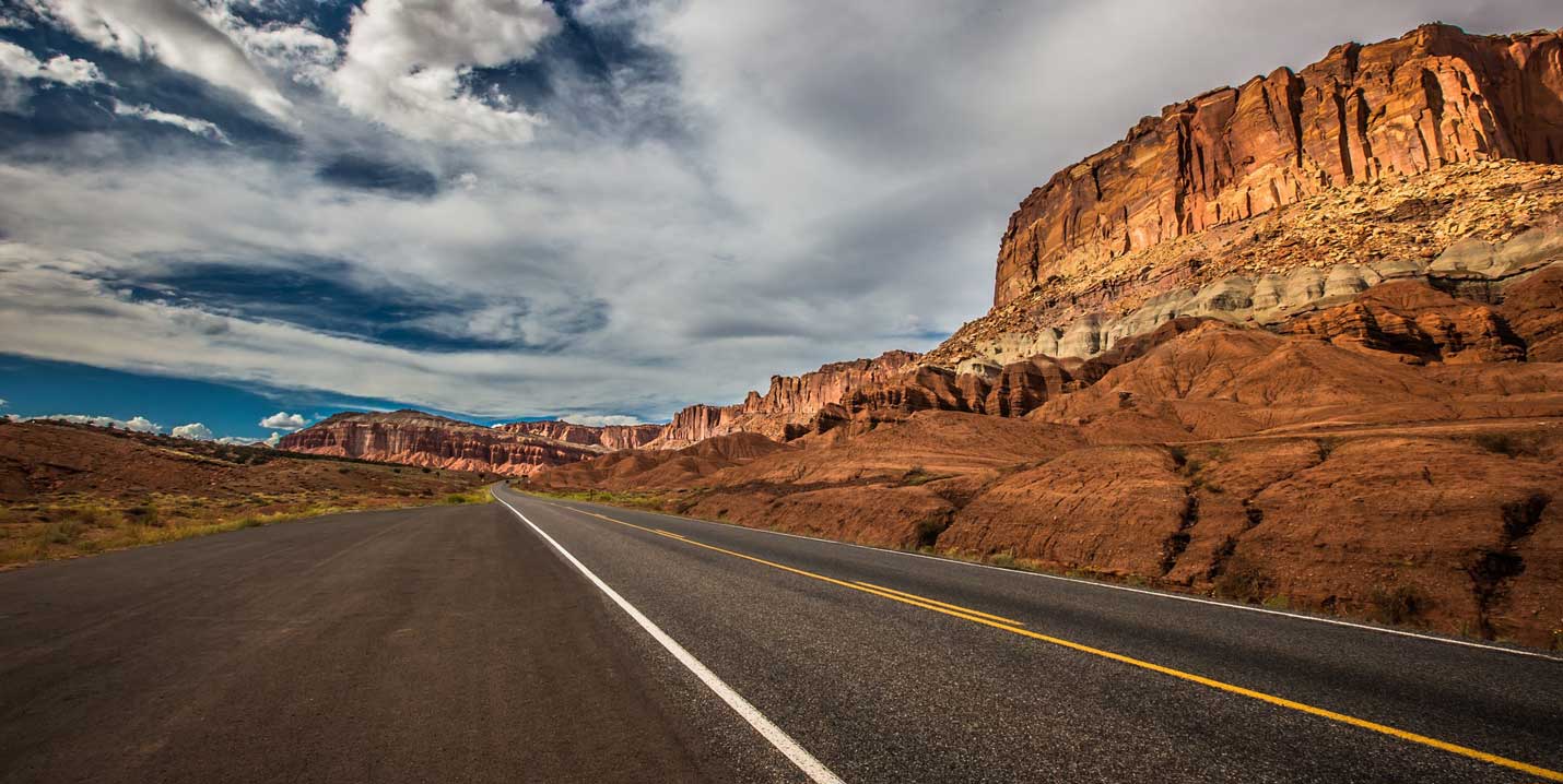 Asphaltierte Straße führt durch eine weite Landschaft mit roten Felsformationen unter bewölktem Himmel