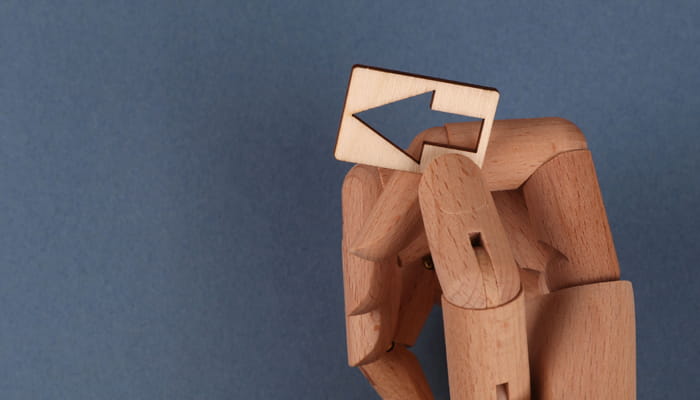 Holzfigur hält ein kleines Schild mit einem nach links gerichteten Pfeil vor den Kopf